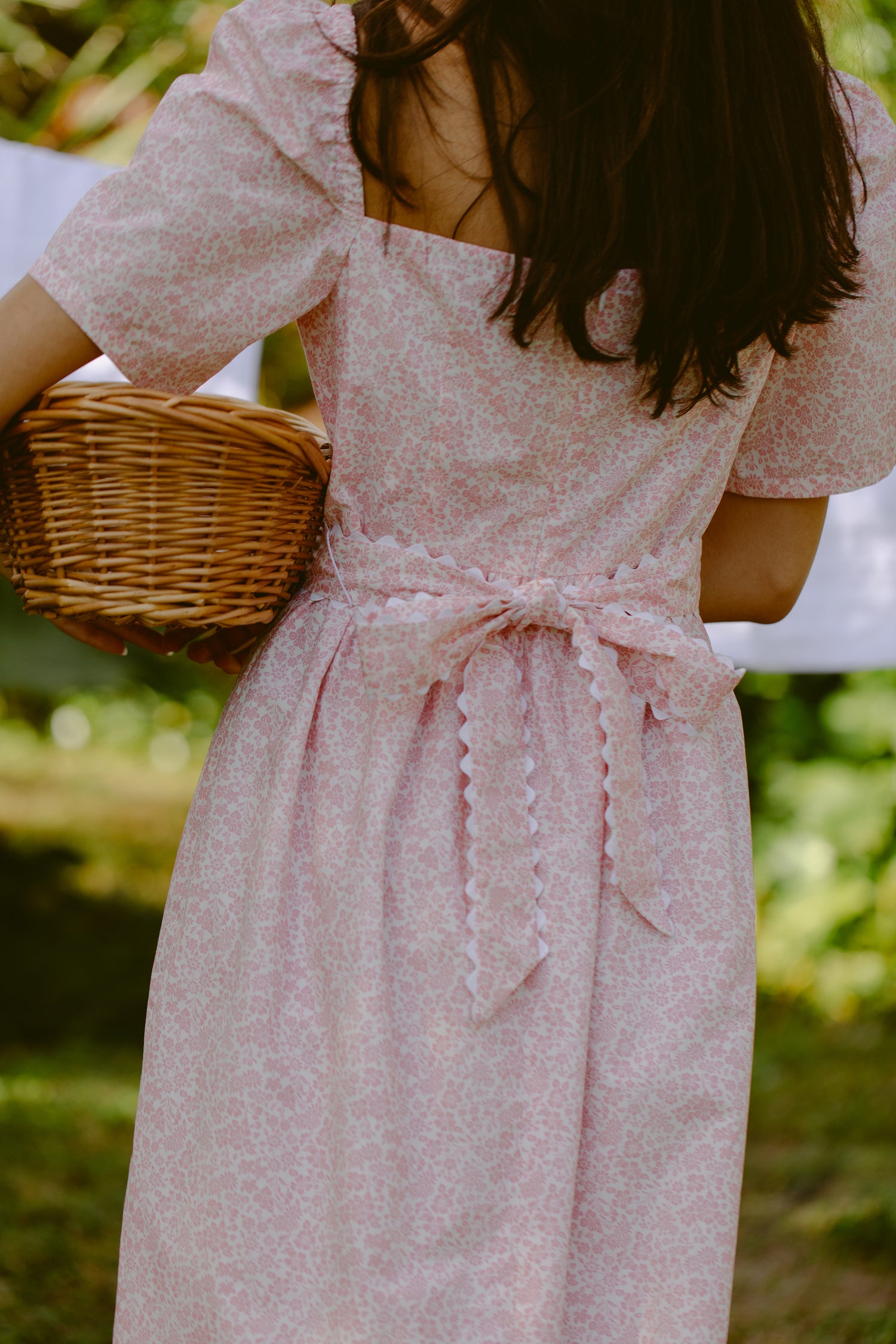 Back view of a woman holding a basket. She is wearing a pink dress with a sash. 