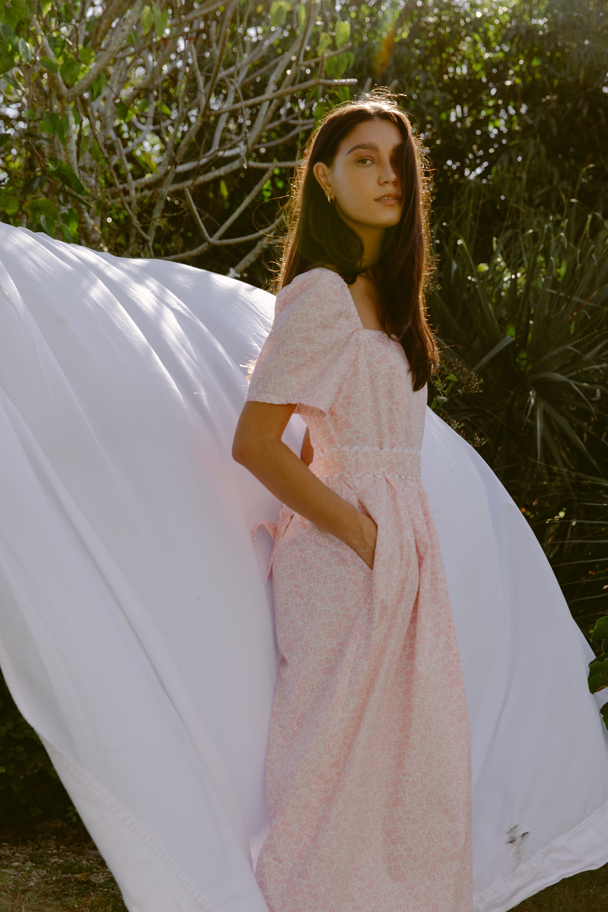 Woman in a pink cotton dress standing in front of a clothing line with a sheet draped over.